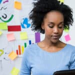 Woman stood in front of a whiteboard looking at a laptop.