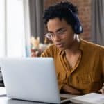 Woman with short curly hair and headphones on sat at a desk making notes in her notepad whilst looking at her laptop.