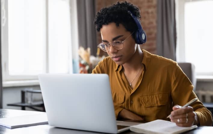 Woman with short curly hair and headphones on sat at a desk making notes in her notepad whilst looking at her laptop.