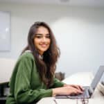 Girl with brown curly hair typing on a laptop and smiling at a camera.