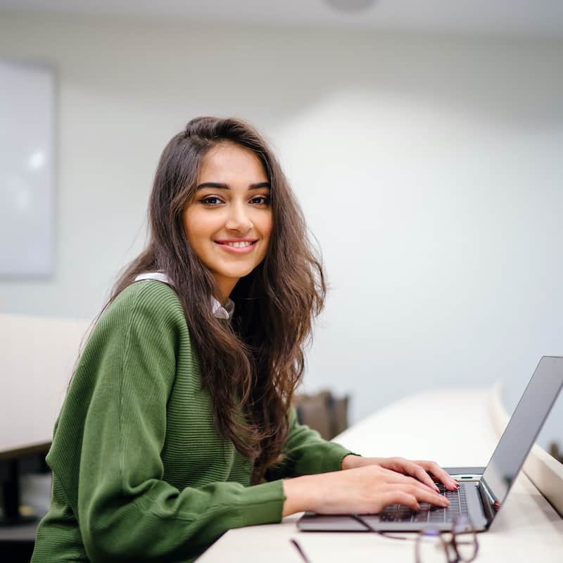 Girl with brown curly hair typing on a laptop and smiling at a camera.