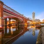 Image of the River Irwell running through Deansgate in Manchester with 3 barges a bridge above and a high rise apartment building in the background.