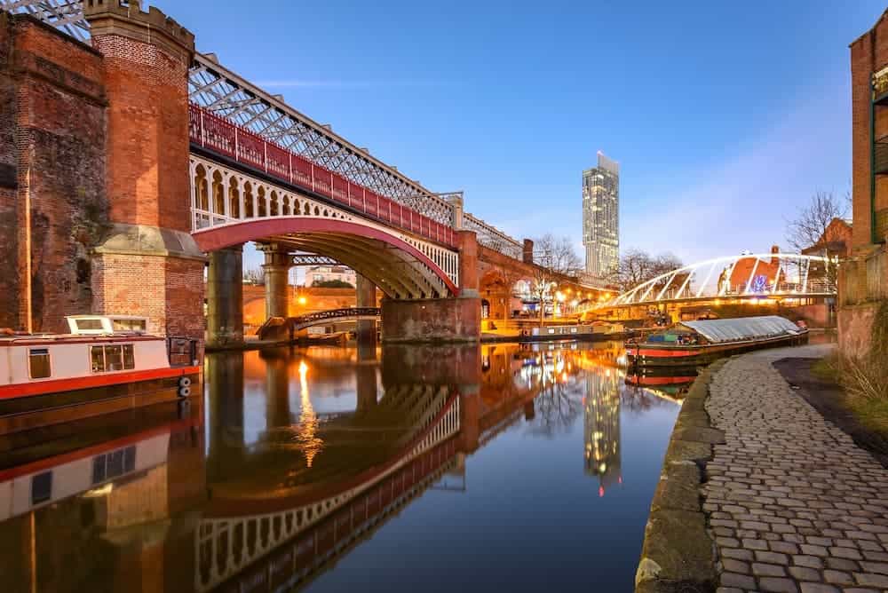 Image of the River Irwell running through Deansgate in Manchester with 3 barges a bridge above and a high rise apartment building in the background.