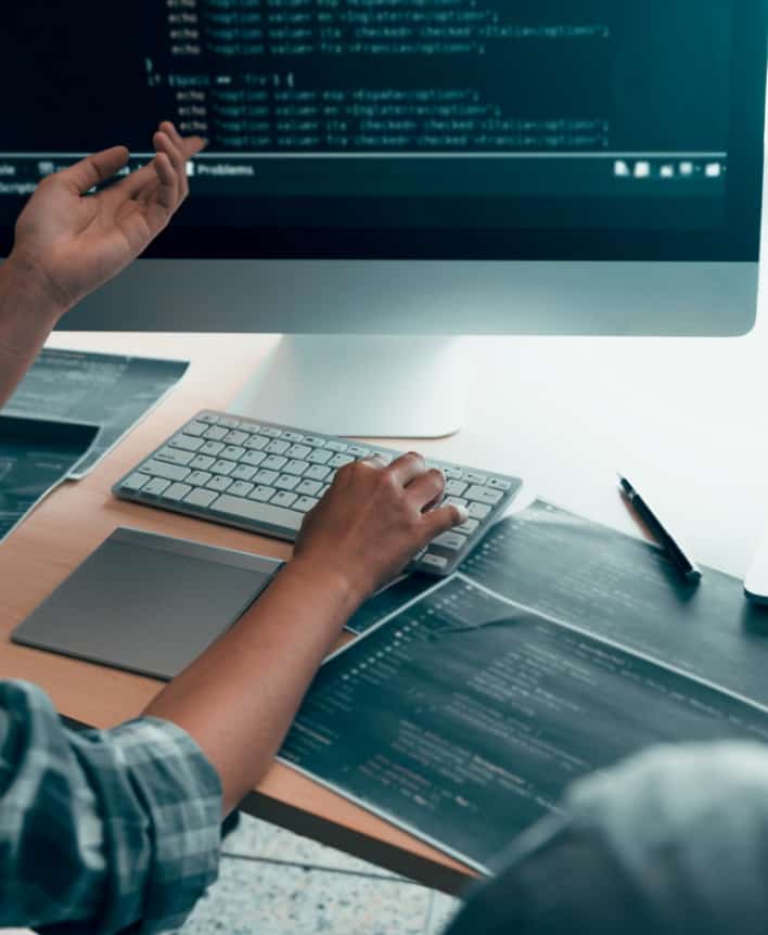 Person sat at a desk typing on a keyboard with their right hand and pointing at the computer screen with their left hand.