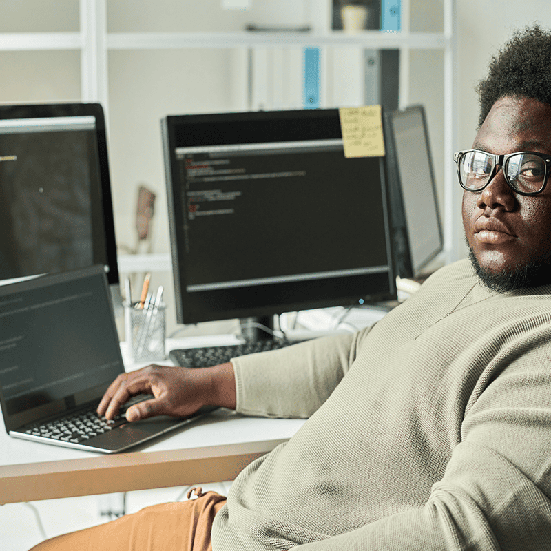 Man wearing glasses looking seriously at the camera with a laptop and monitor displaying code.