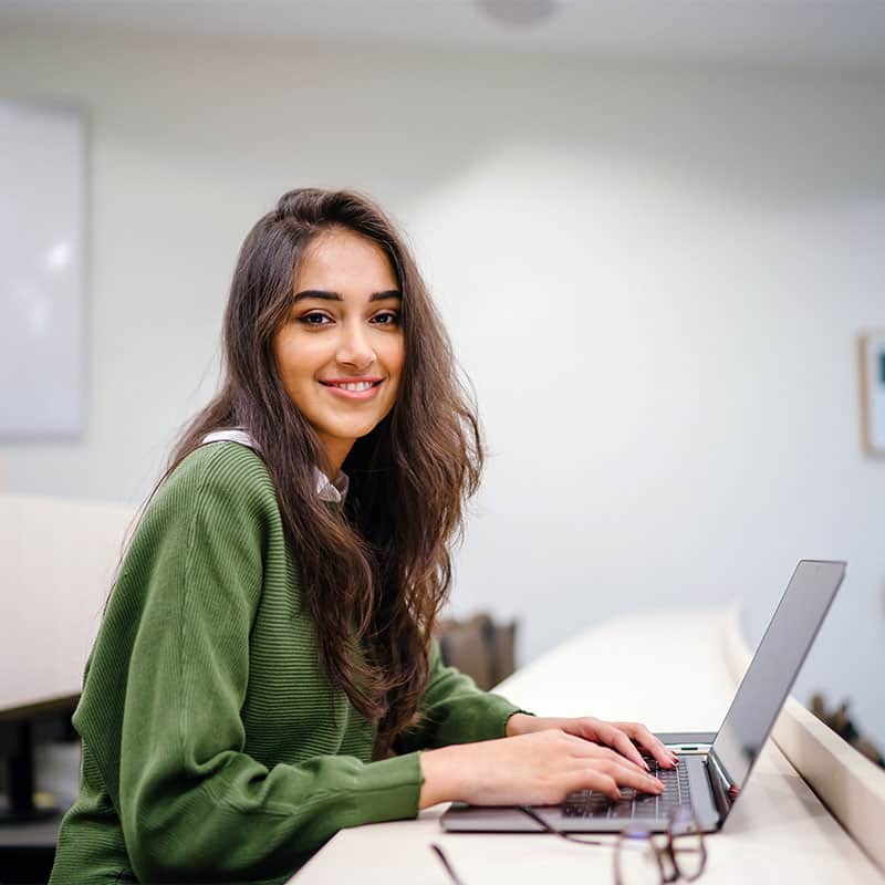 Girl with long wavy dark brown hair typing on a laptop smiling at the camera