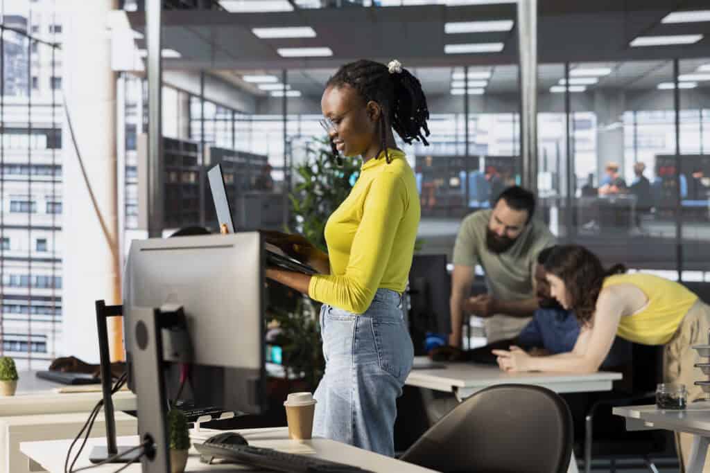 Woman typing on laptop keyboard next to staff members in modern workplace